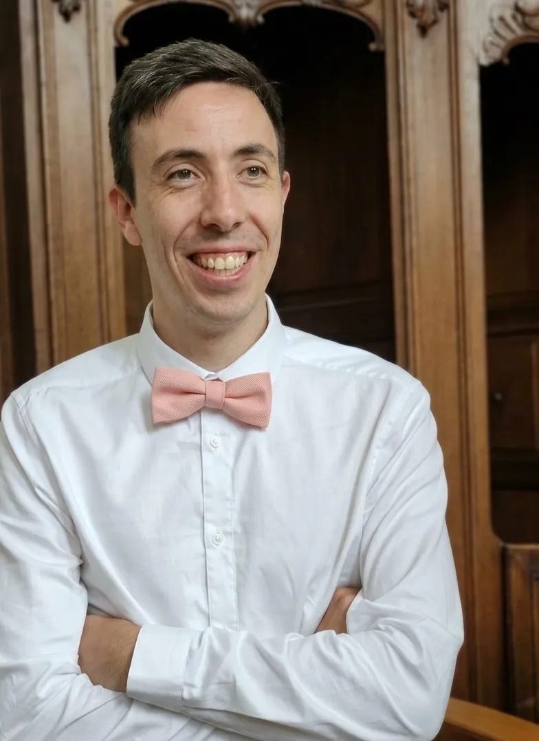 Martin Marot, souriant, bras croisés, en chemise blanche et nœud papillon rose.